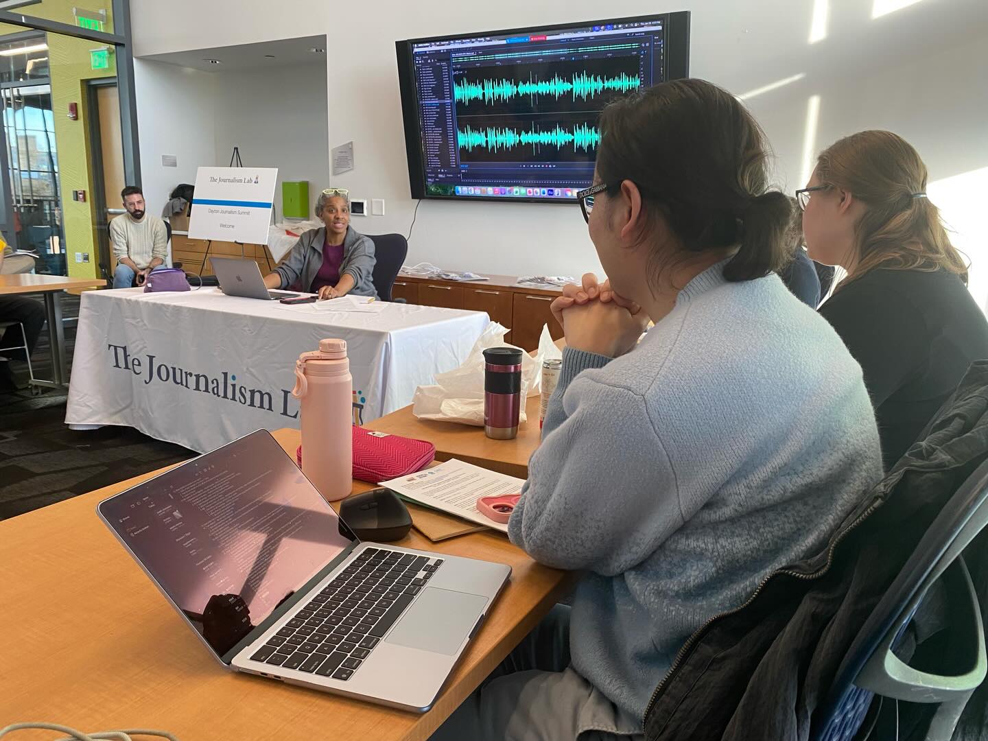 Kathryn Mobley conducts a workshop at a table with The Journalism Lab banner, with participants watching an audio editing demonstration on a large screen at the Downtown Dayton Metro Library Branch